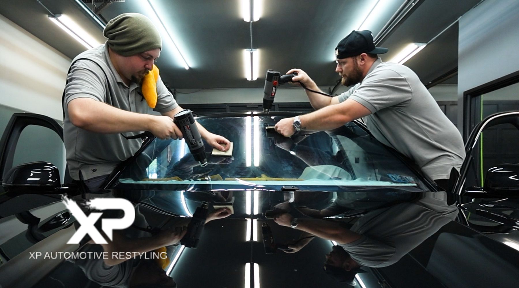 Two technicians work on installing a window tint on a car windshield in a well-lit garage. They are using heat guns and smoothing tools to ensure a precise application. The logo "XP Automotive Restyling" is displayed prominently in the lower left corner of the image.