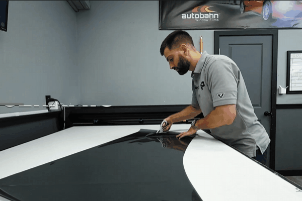 A technician is carefully cutting a large sheet of ceramic window tint film on a workbench, preparing it for installation. The background shows an "Autobahn Window Films" banner, emphasizing the premium quality of the tint. The process highlights precision and attention to detail in automotive restyling.