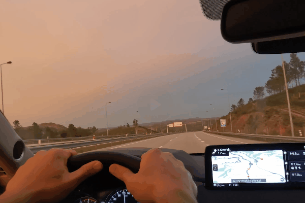 A driver grips the steering wheel while driving on a mostly empty highway during a sunset or early evening, with a GPS navigation screen visible on the dashboard. The tinted windows reduce glare from the fading sunlight, offering a clearer and more comfortable view of the road ahead.