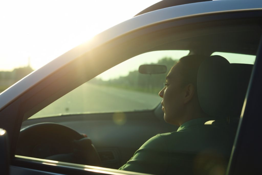 A person is driving a car during sunset, with the bright sunlight shining through the driver's side window. The scene emphasizes the potential discomfort caused by direct sun exposure, highlighting the benefits of window tint for reducing glare and enhancing comfort while driving.