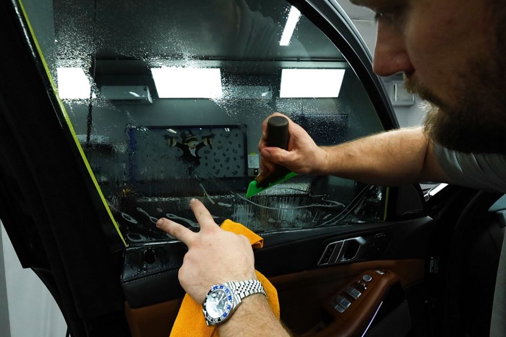 A close-up of a technician meticulously applying window tint to a car door window using a squeegee tool. The window is wet, and the technician uses a cloth to wipe away excess water while ensuring a smooth and bubble-free tint installation. This image highlights the precision and care taken during the tinting process.