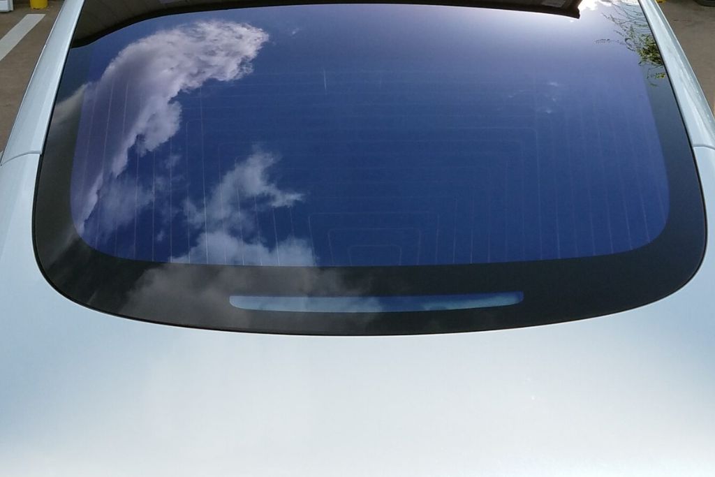 Rear window of a car with a crystalline window tint, reflecting a blue sky with scattered clouds. The tint appears light yet effectively reduces glare while maintaining clear visibility.