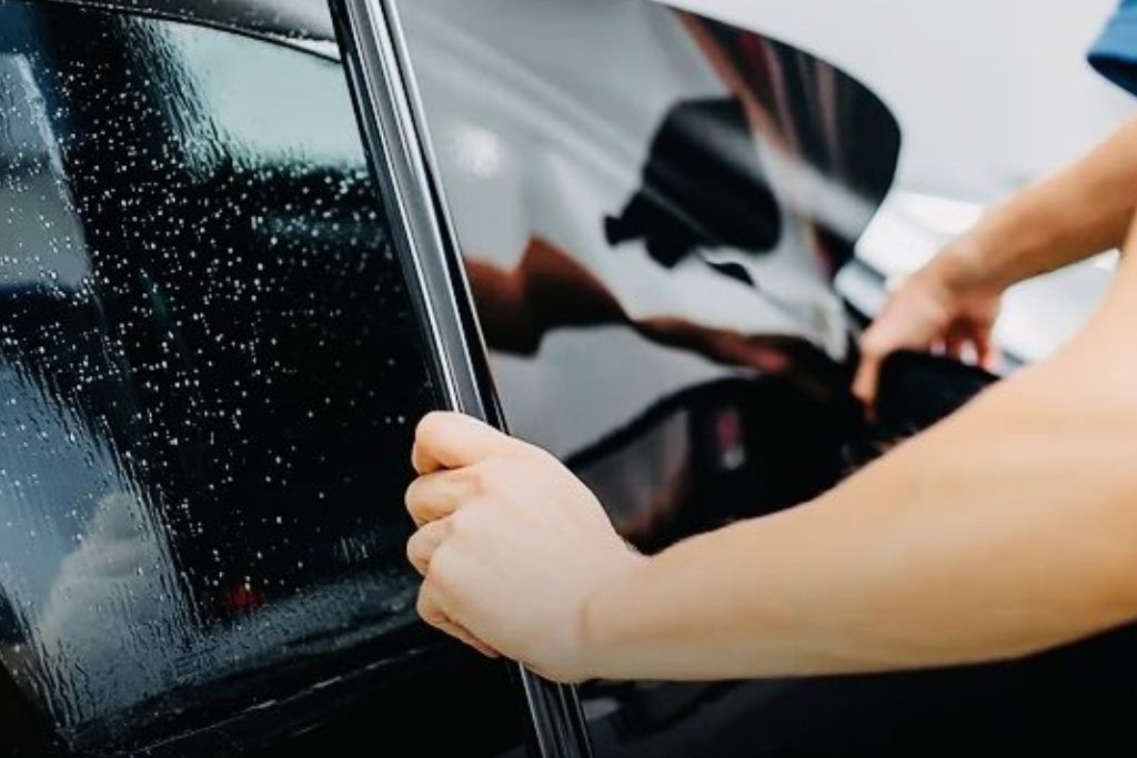 Hands applying a metalized tint film to a car window, with water droplets visible on the glass surface during the installation process. The metalized tint helps reflect heat and UV rays, providing better cooling.