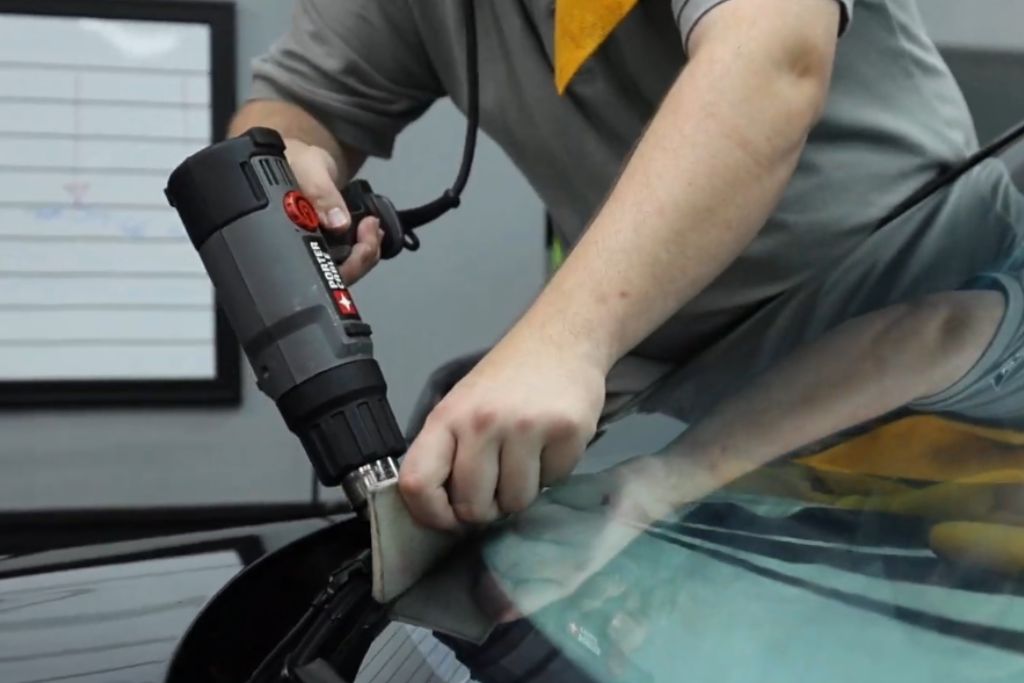 A technician carefully applies heat with a heat gun while smoothing a sheet of window tint onto a car's windshield, ensuring a seamless finish during installation.