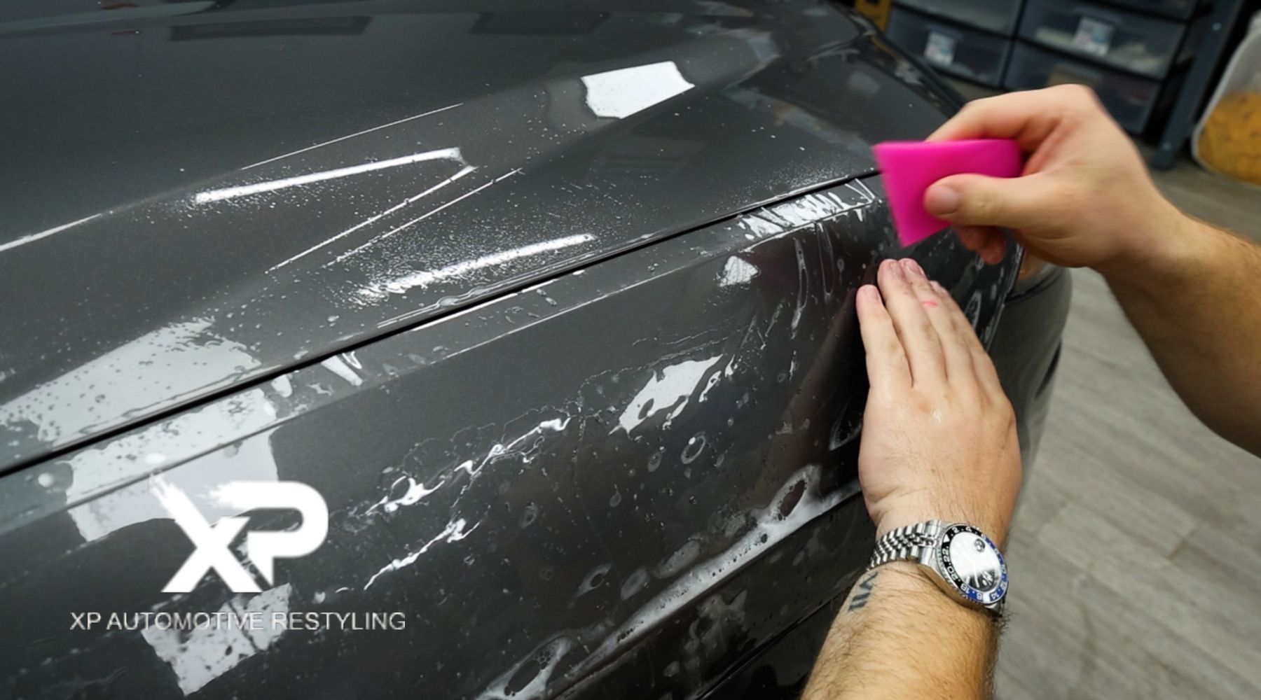 A person applying a clear paint protection film to the hood of a dark gray car using a pink squeegee tool. The surface is wet, with visible water streaks and bubbles being smoothed out. The XP Automotive Restyling logo is displayed in the bottom-left corner.