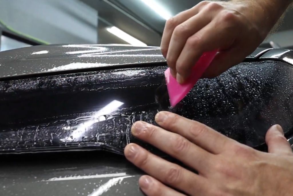 Close-up of hands applying paint protection film to a car's surface using a pink squeegee, with water droplets visible, demonstrating the installation process for gloss or matte finishes.