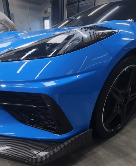 A vibrant blue sports car with a ceramic-coated finish glistens under indoor lighting, showcasing its flawless and reflective surface. A person’s hand is partially visible in the background performing detailing work.