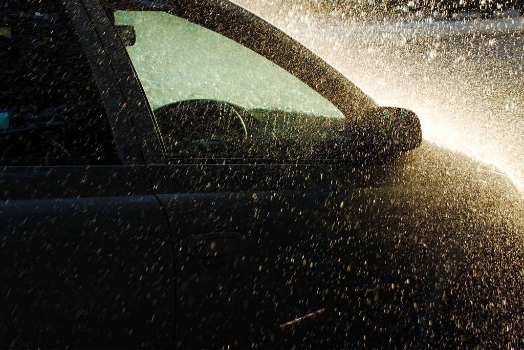 A car being sprayed with water, highlighting how seasonal maintenance protects against elements like road salt and UV rays. The water spray is illuminated by sunlight, creating a dynamic effect on the vehicle's surface.