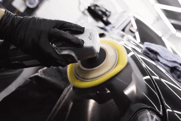 A close-up of a car's glossy black surface being polished with a buffer machine equipped with a yellow pad. A person wearing black gloves carefully operates the machine, ensuring a smooth and shiny finish.