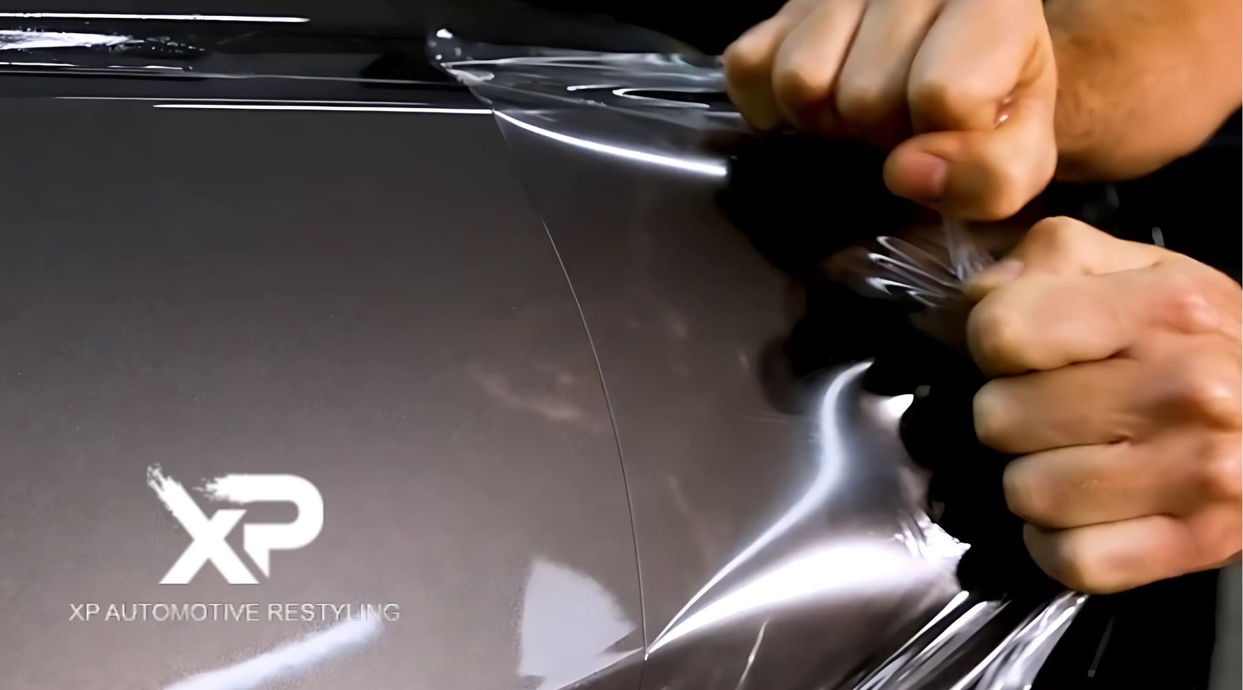 Close-up of a person’s hands peeling off a clear protective film from a car’s glossy surface. The image highlights the removal process of a paint protection film (PPF), revealing the car’s pristine finish underneath. The "XP Automotive Restyling" logo is visible in the bottom left corner.