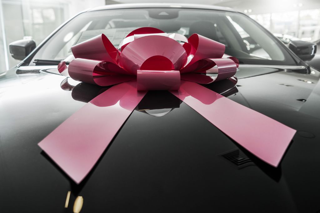 A brand-new black car in a showroom with a large pink bow placed on the hood, symbolizing a gift or special purchase. The car’s glossy paint reflects the bright lights, emphasizing its pristine condition.