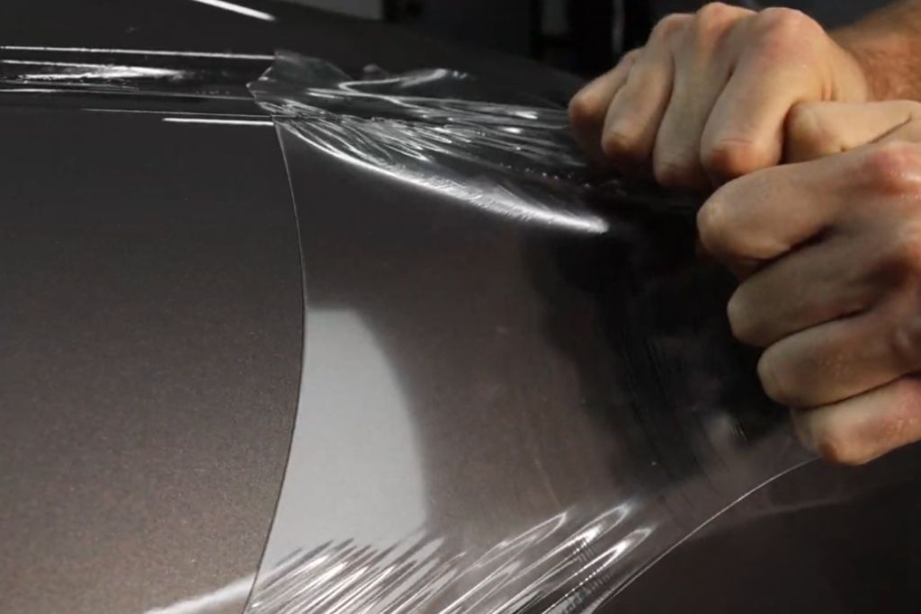 A close-up of a person forcefully peeling off a clear paint protection film (PPF) from a car’s metallic surface. The transparent film is being stretched and removed, revealing the glossy paint underneath. The image highlights the durability and removability of PPF, which helps protect the car’s finish from scratches and environmental damage.