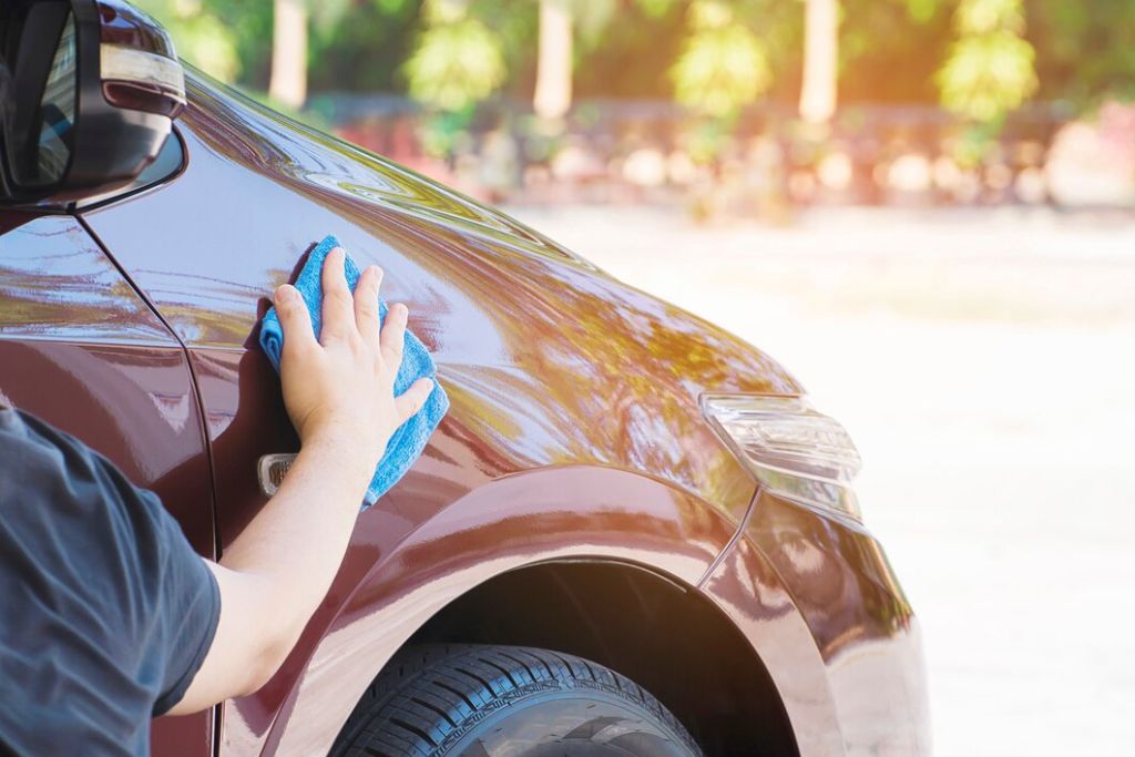 A person wiping down the glossy exterior of a brown car with a blue microfiber cloth. The sunlight reflects off the clean surface, emphasizing the well-maintained finish. The background features a bright outdoor setting with trees and soft-focus greenery, highlighting the car’s polished look.