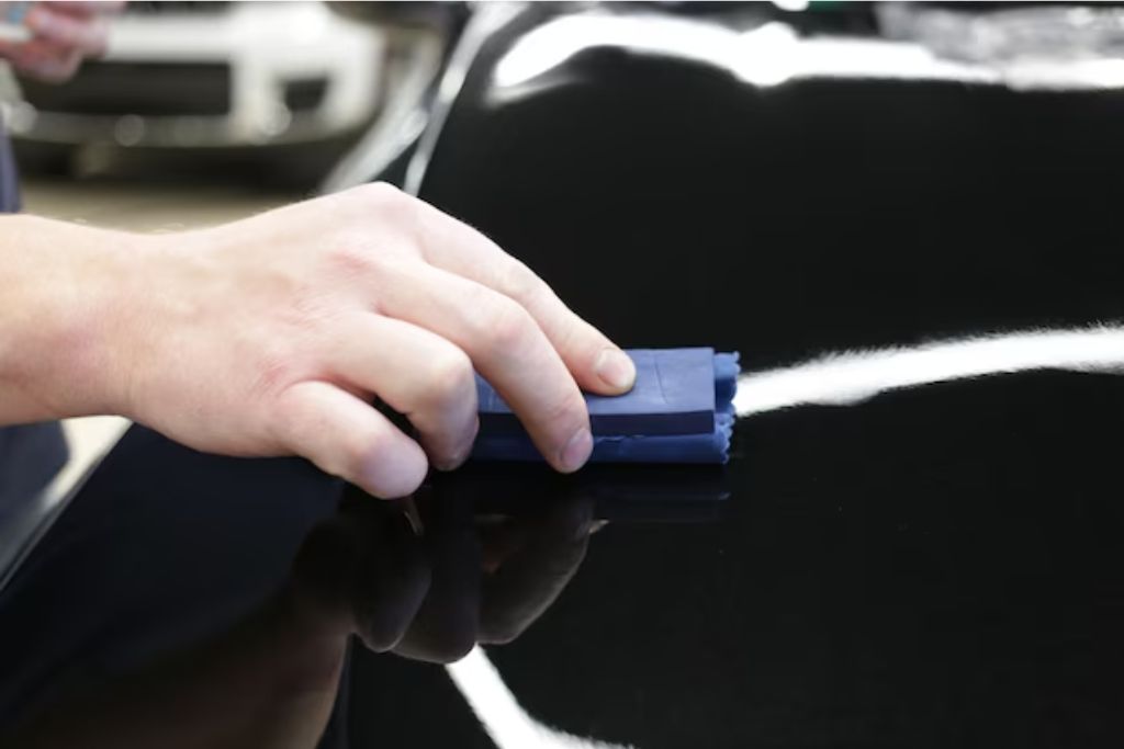 A close-up of a hand applying ceramic coating to a glossy black car surface using a foam applicator wrapped in a microfiber cloth. The coating creates a sleek, reflective finish, enhancing the vehicle's shine and adding an extra layer of protection over the paint or PPF. A blurred background of a garage or detailing shop is visible.