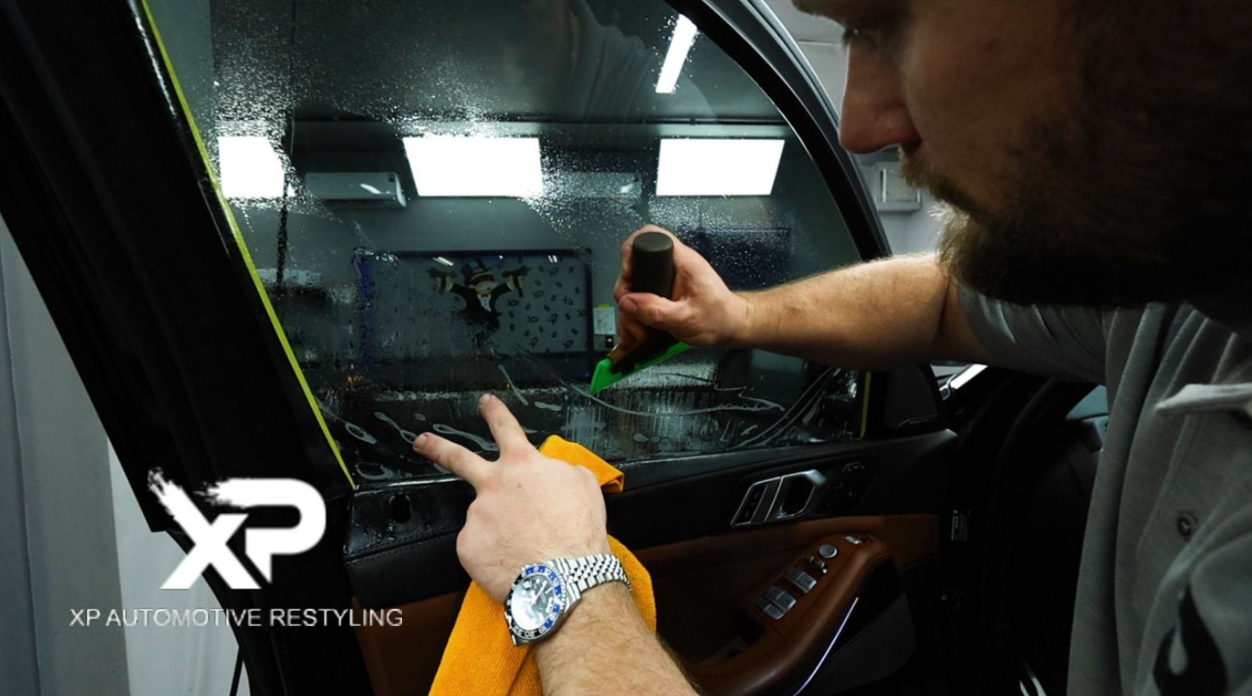 A professional installer carefully applies a privacy window tint to a car’s side window using a green squeegee tool. The window is wet, and the reflection of the workspace is visible. The "XP Automotive Restyling" logo is displayed in the corner.