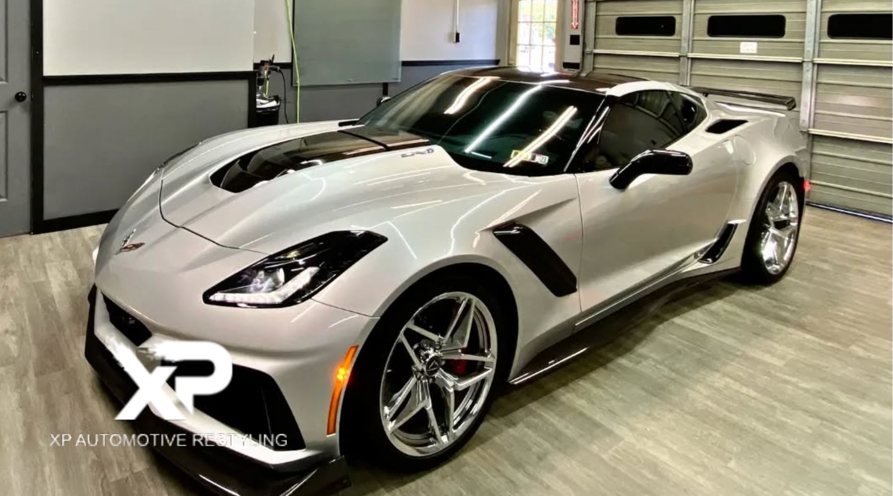 A sleek white Chevrolet Corvette ZR1 with tinted windows and glossy paint sits inside a professional detailing garage with modern lighting and wood flooring. The "XP Xpress Window Tinting" logo is visible in the lower left corner, indicating a high-quality automotive service location.