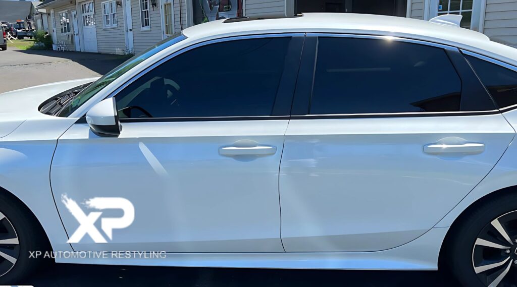 A sleek white sedan with dark-tinted windows parked outside a shop. The deep window tint enhances privacy, reduces glare, and blocks UV rays, improving both comfort and interior protection. The "XP Automotive Restyling" logo is visible in the bottom left corner, highlighting the tinting service provider.