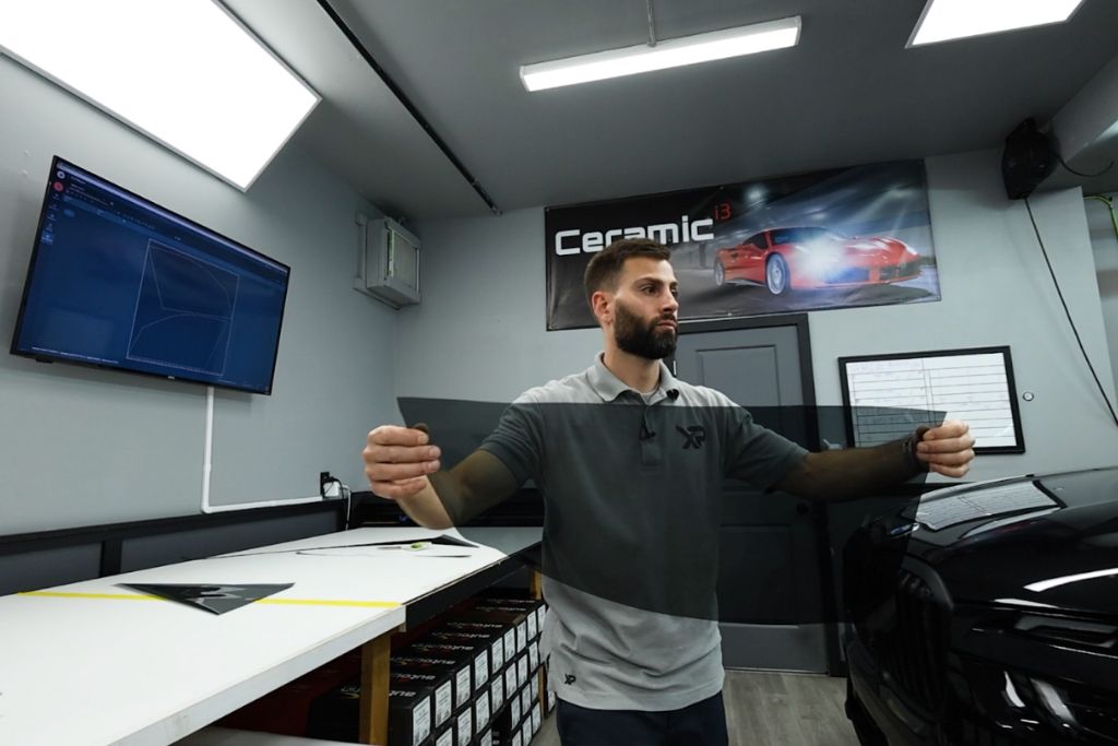 A professional installer at XP Automotive Restyling holds up a pre-cut window tint film inside a modern detailing shop. A "Ceramic" banner and digital cutting software display are visible in the background.