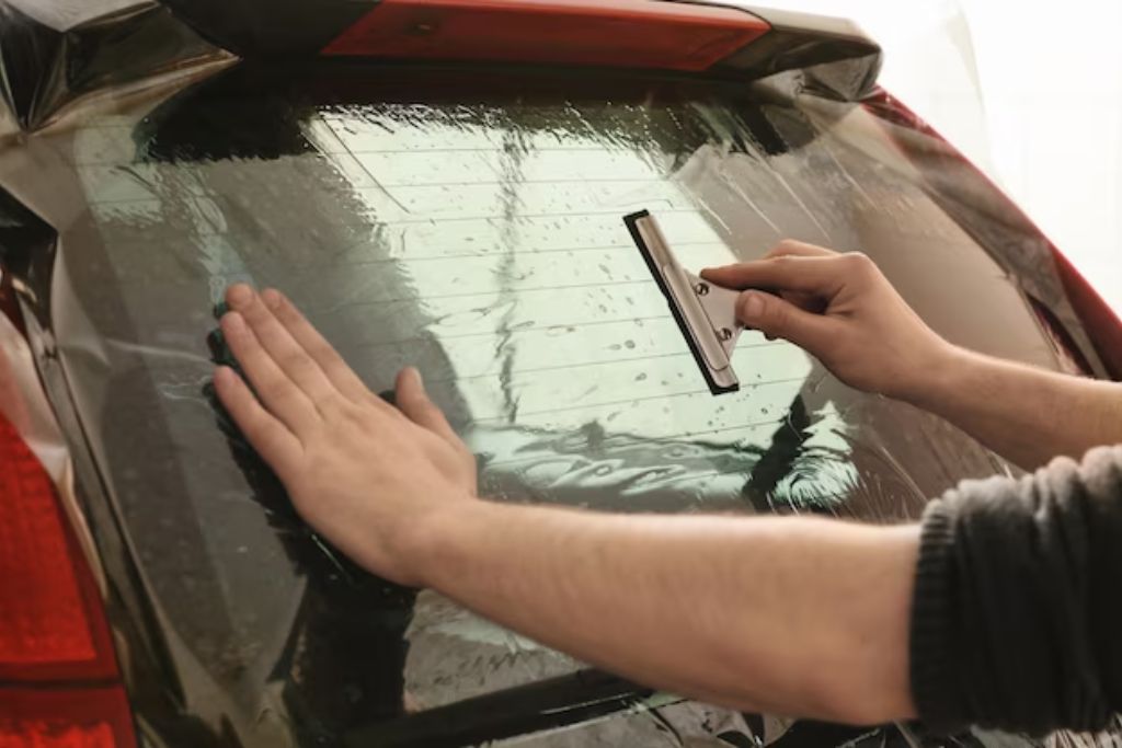 A person installs a window tint on the rear windshield of a vehicle, using a squeegee to smooth out the film while holding it in place with their other hand. The wet application process is visible.