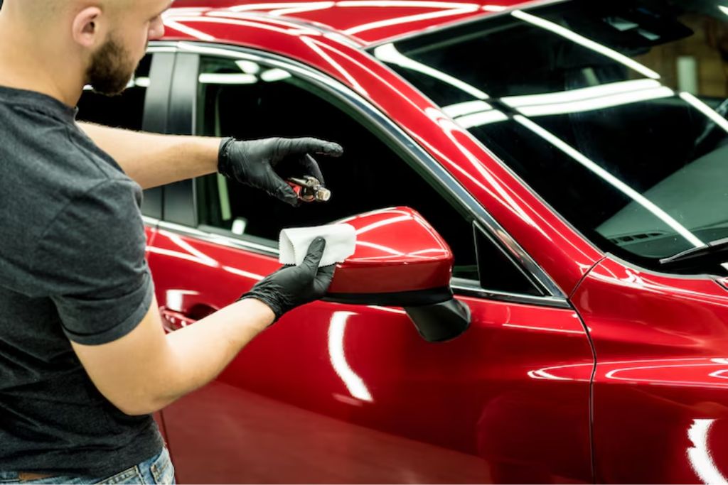 Man wearing black gloves applying ceramic coating with an applicator pad to the red side mirror of a shiny car, highlighting the careful detailing process.