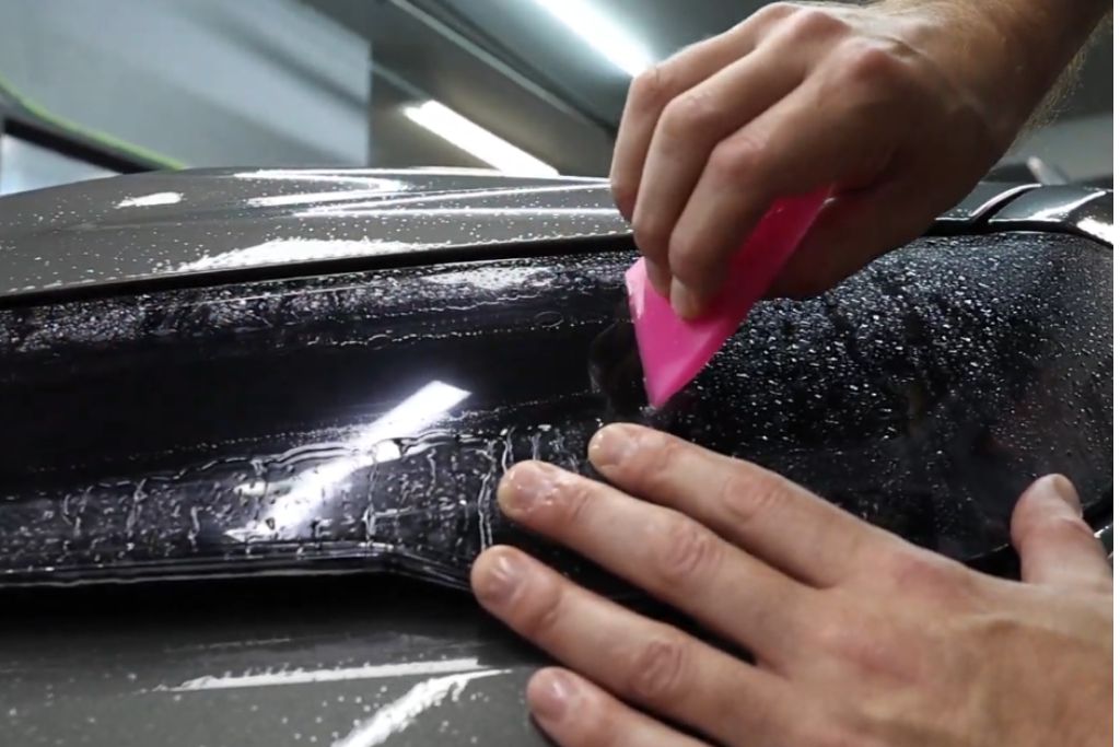 Close-up of hands applying paint protection film to a wet car surface using a pink squeegee, with visible water droplets and reflections on the dark glossy finish.
