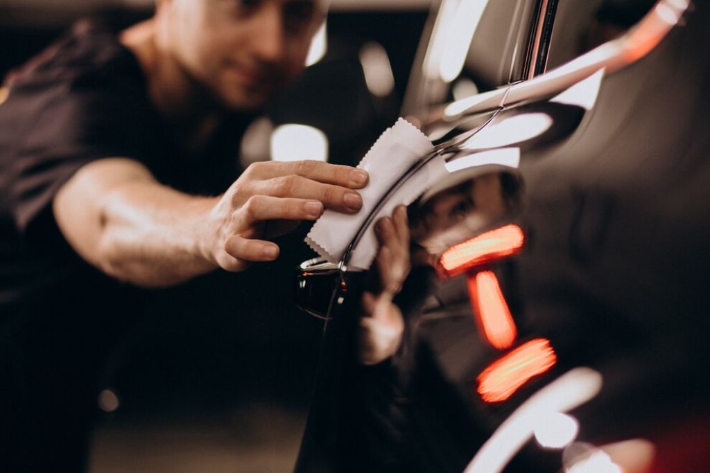 Man carefully applying ceramic coating to a glossy black car door using a white cloth, with a focused reflection showing the detailing precision under bright lights.