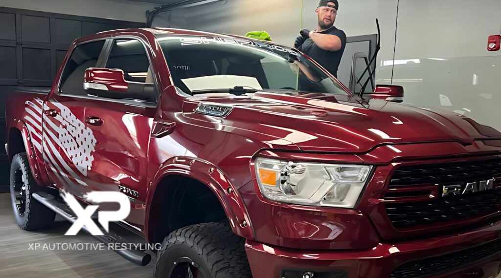 A man applies ceramic coating to a shiny, deep red RAM 1500 truck with black wheels and an American flag graphic on the side, inside a clean garage at XP Automotive Restyling.