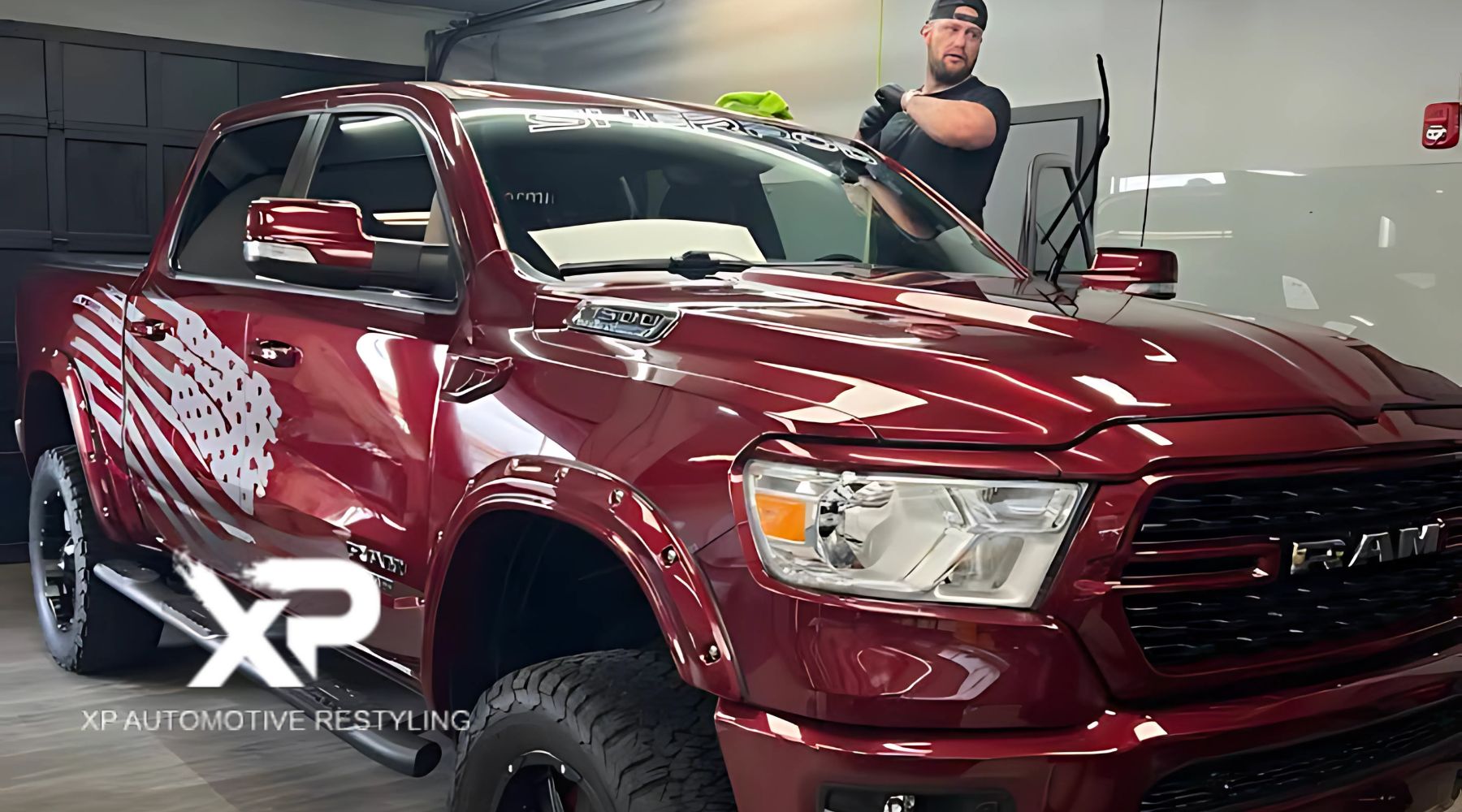 A man applies ceramic coating to a shiny, deep red RAM 1500 truck with black wheels and an American flag graphic on the side, inside a clean garage at XP Automotive Restyling.