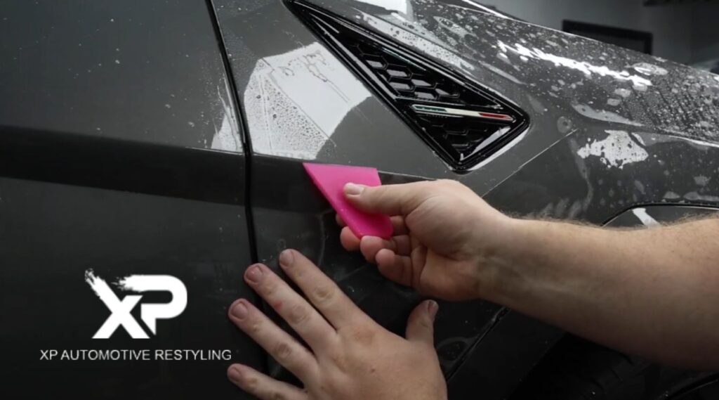 A person applies paint protection film to a glossy dark car using a pink squeegee, smoothing the film around the panel edge near the vent. Water droplets and soap residue are visible on the surface, with the "XP Automotive Restyling" logo in the lower left corner.
