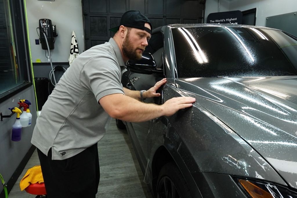 A technician in a gray XP Automotive Restyling shirt carefully smooths paint protection film on a dark gray SUV inside a detailing shop, with water droplets visible on the hood and fender.