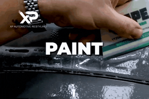 A close-up of a technician applying paint protection film with a squeegee tool on a wet car surface. Bold white text reading “PAINT” appears across the center of the screen during the application.