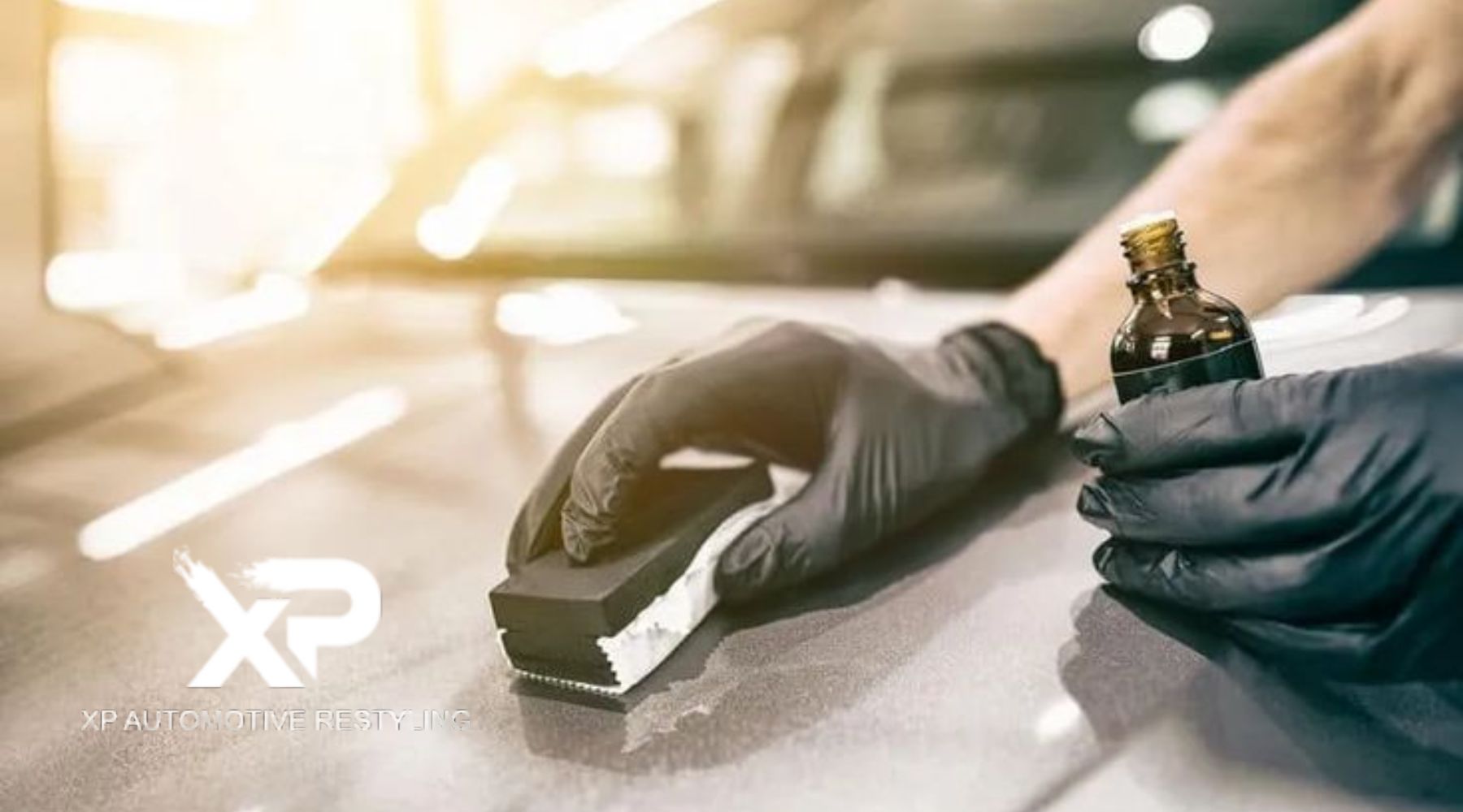 Close-up of a person wearing black gloves applying ceramic coating to a car’s surface using an applicator pad and a small amber bottle, with sunlight reflecting off the glossy paint. The XP Automotive Restyling logo appears on the bottom left corner.