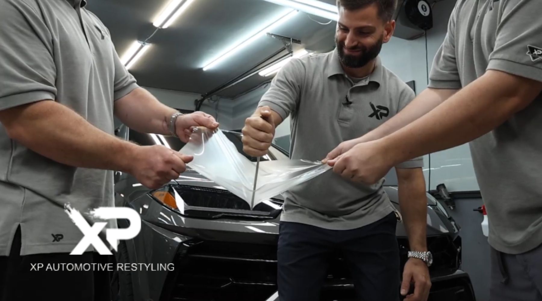 Three men wearing matching grey XP Automotive Restyling shirts demonstrate the strength of a transparent paint protection film by stretching it and stabbing it with a metal tool in front of a dark-colored car inside a well-lit garage. The XP Automotive Restyling logo is visible in the bottom left corner.