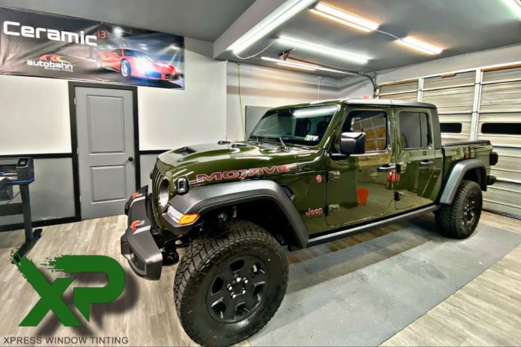 A glossy olive green Jeep Gladiator Mojave is parked inside a clean detailing bay at XP Automotive Restyling, featuring window tint and a likely PPF application. A large banner for Autobahn Ceramic window film is mounted on the wall, and the XP Xpress Window Tinting logo is visible in the foreground.