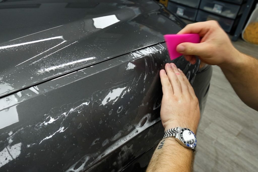 Close-up of a person using a pink squeegee tool to apply paint protection film to the fender of a dark gray vehicle. The film is being smoothed over a wet surface, with visible water and air bubbles during installation.