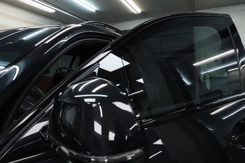 Close-up of a darkly tinted car window in a garage, reflecting overhead lights and concealing the interior—highlighting the privacy and security benefits of automotive window tint.