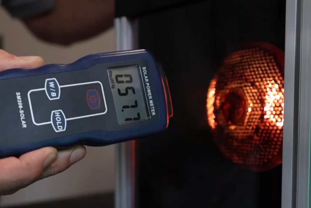 Close-up of a handheld solar power meter reading 057.7 in front of a glowing heat lamp, demonstrating how window tint blocks infrared heat to protect vehicle interiors.