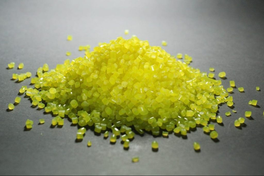 A close-up photo of a small mound of translucent yellow resin pellets scattered on a dark surface, representing the raw materials used in ceramic coating formulations for binding and adhesion.
