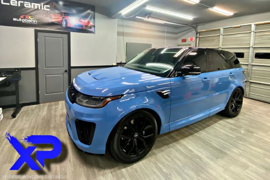 A glossy blue Range Rover SUV with tinted windows sits in a well-lit garage with XP branding on the floor and a banner reading "Ceramic" on the wall. The vehicle's reflective surface showcases the smooth, high-gloss finish typical of ceramic coatings.
