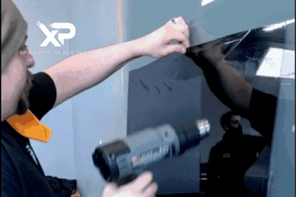 A technician at XP Automotive Restyling uses a heat gun and squeegee to precisely apply window tint film to a vehicle, showcasing expert installation and attention to detail.