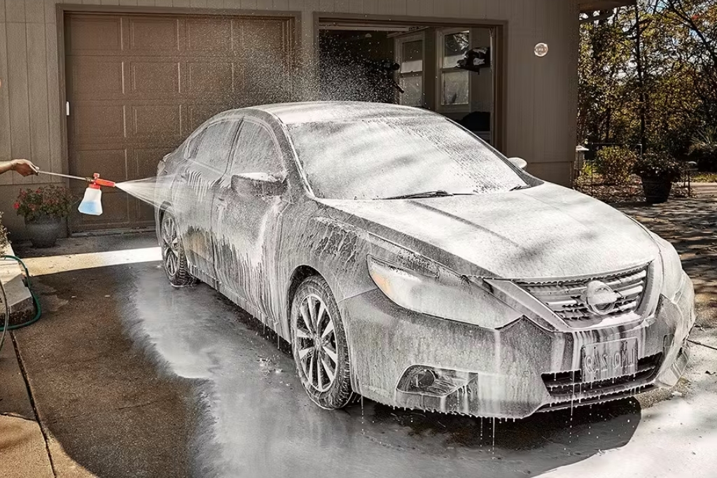 Person spraying a thick layer of foam onto a sedan in a driveway using a foam cannon, illustrating proper hand-wash maintenance for a ceramic-coated vehicle to preserve protection against chemicals and bird droppings.