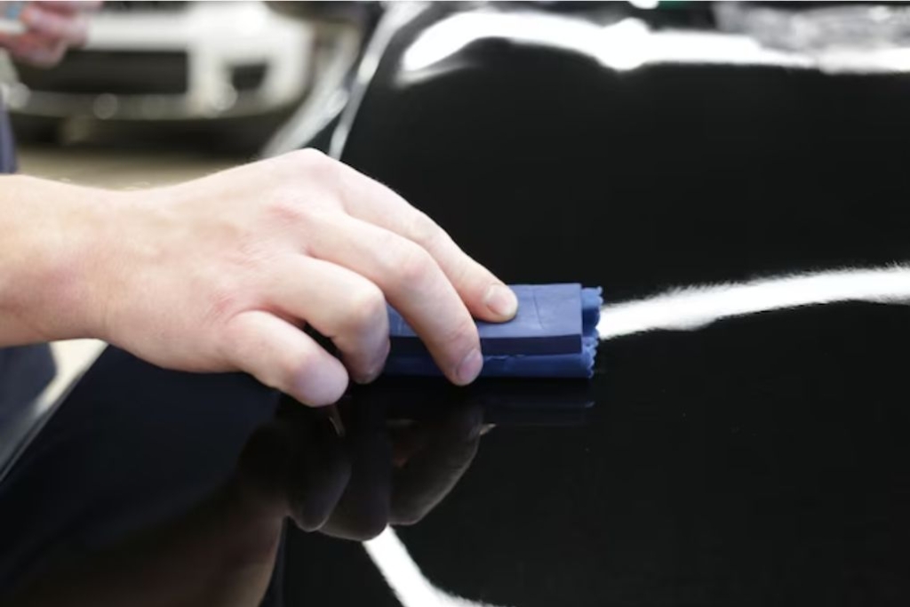 A person’s hand glides a clay bar across a glossy black car surface to remove contaminants before reapplying ceramic coating. Proper surface prep like this is essential when considering can you apply a new coating over the old one.