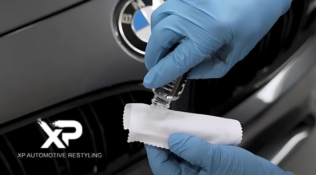 Close-up of a person wearing blue gloves pouring ceramic coating onto a white applicator cloth in front of a BMW grille, with the XP Automotive Restyling logo in the corner. This hands-on visual demonstrates the process relevant to the question Does Ceramic Coating Need to Be Reapplied.