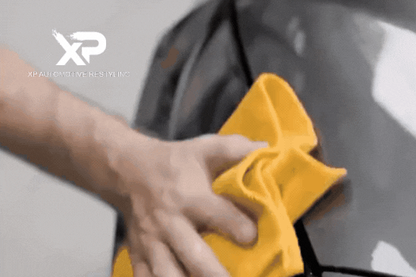 Close-up of a hand using a yellow microfiber cloth to buff a glossy black car surface, showcasing the final stage of ceramic coating application. The XP Automotive Restyling logo appears in the corner, reinforcing the message that expert application matters for lasting results.