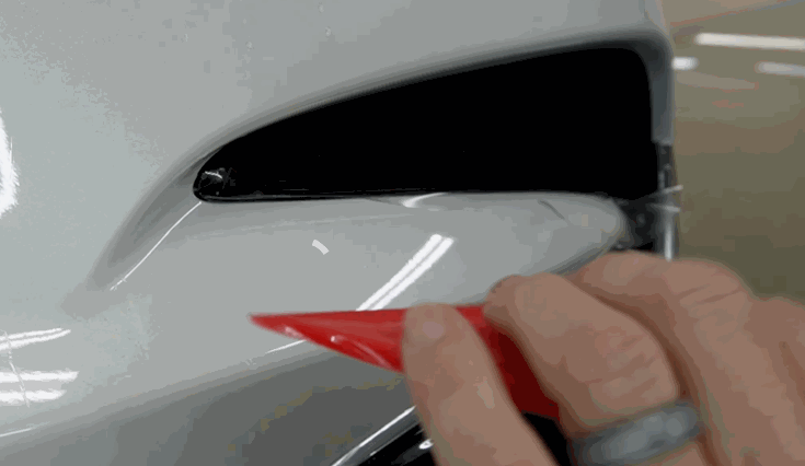 Close-up of a red squeegee tool being used to tuck paint protection film into the corner edge of a white bumper.