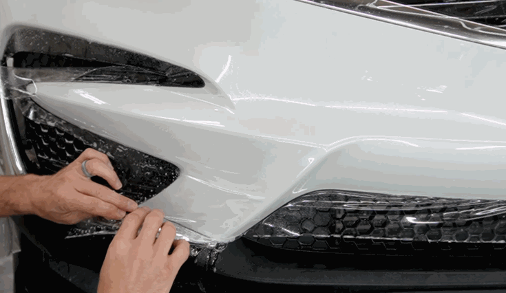 A technician smooths paint protection film along the front bumper of a white car, pressing it into the body lines and around the headlights.