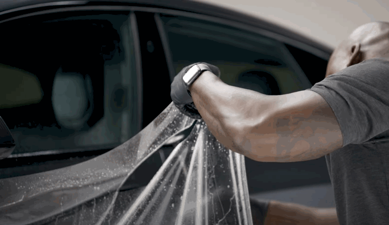 A man in a gray shirt and gloves pulls a wet sheet of paint protection film across a car door during installation.