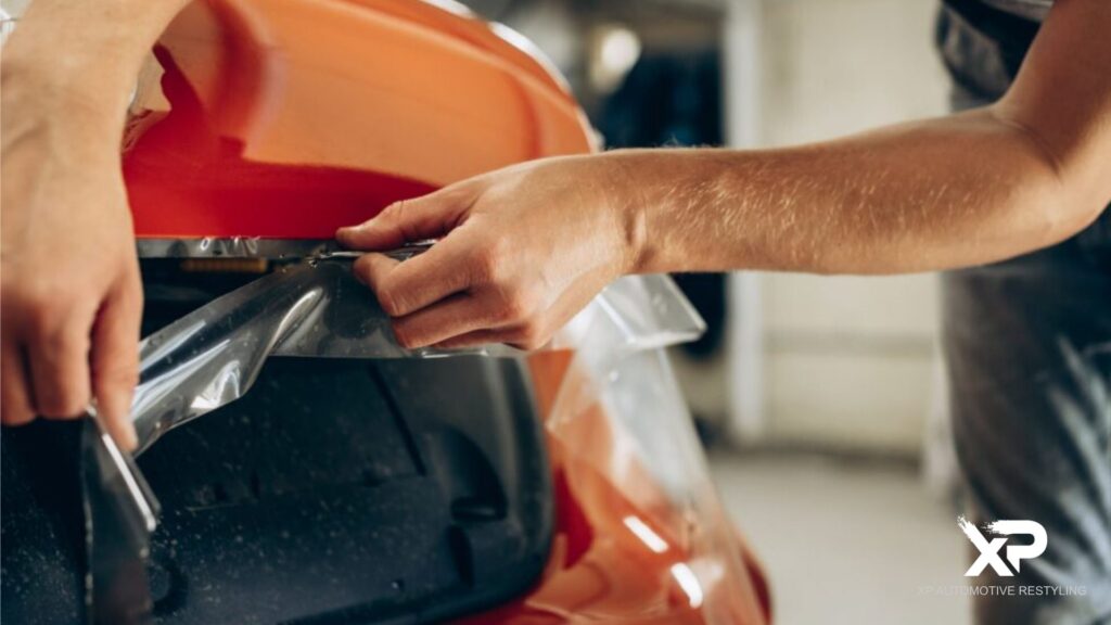 Hands applying and trimming clear paint protection film to the front of a shiny red sports car, with XP Automotive Restyling logo in the corner.