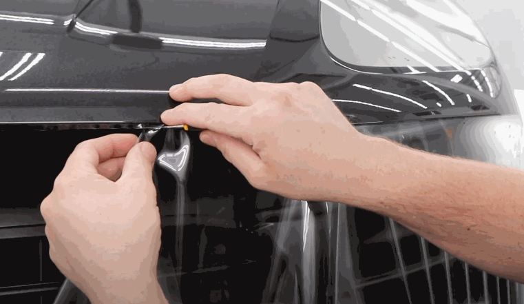 Installer uses a squeegee tool to smooth a paint protection film over the front bumper of a black car, trimming near the logo.