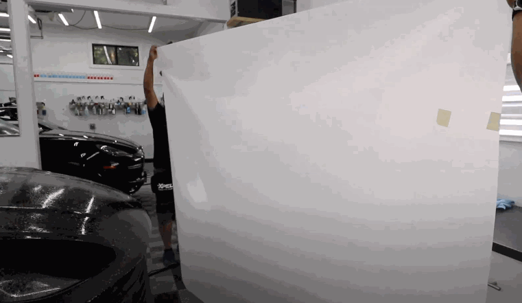 Two installers lift and position a large sheet of paint protection film inside a detailing workshop with black cars in the background.
